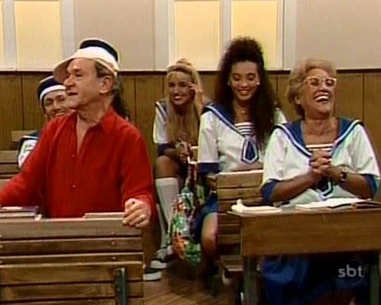 Cena da Escolinha do Golias: Ronald Golias em pé ao lado de uma carteira escolar, usando camisa vermelha e chapéu branco; Nair Bello está sentada ao lado, rindo. Ao fundo, outros personagens vestem uniformes de marinheiro.