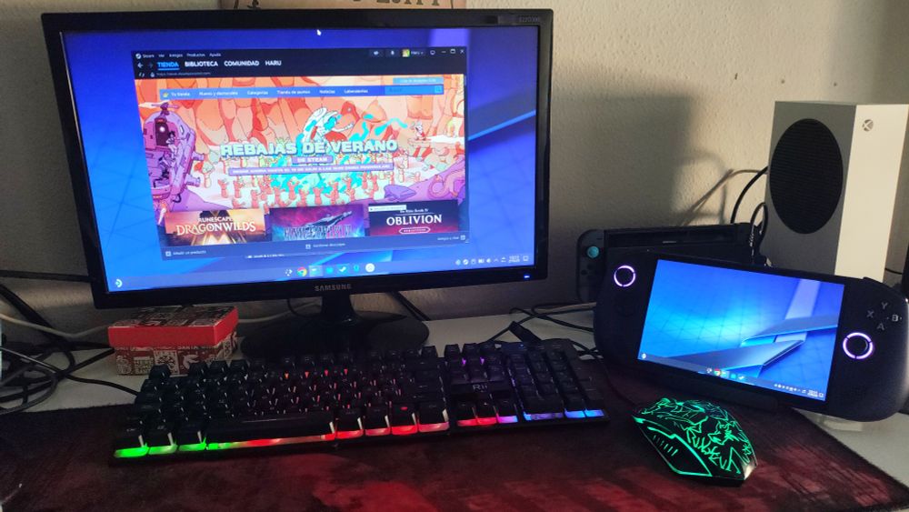Ordenador de escritorio usando la legion go s como pc y segunda pantalla, aparecen tambien el monitor con steam abierto, un teclado y un raton con luces rgb sobre una alfombrilla de escritorio roja y negra que se extiende por el escritorio.

De fondo adicionalmente se aprecian cables, una caja roja, la switch y la xbox series s