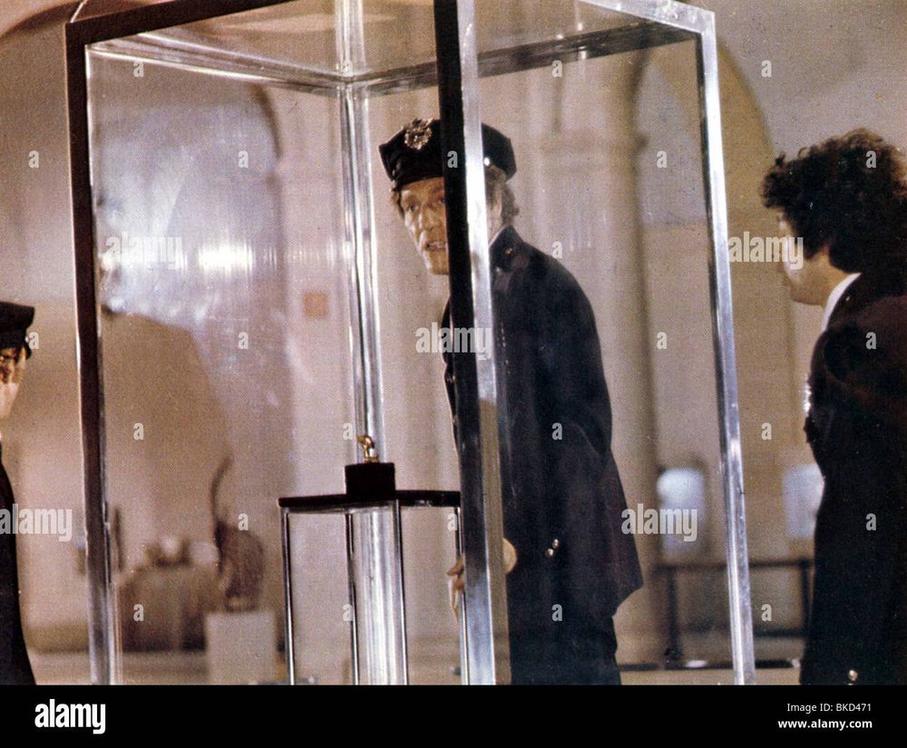 Movie still showing a man in a police uniform standing inside a tall glass case, next to a pedestal holding a small object. Two men also in police uniforms stand outside the case on either side of it.