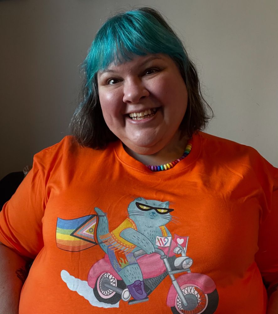 Fat babe Joanna in a bright orange t-shirt featuring a cartoon fat cat on a pink motorcycle, flying the progressive pride flag