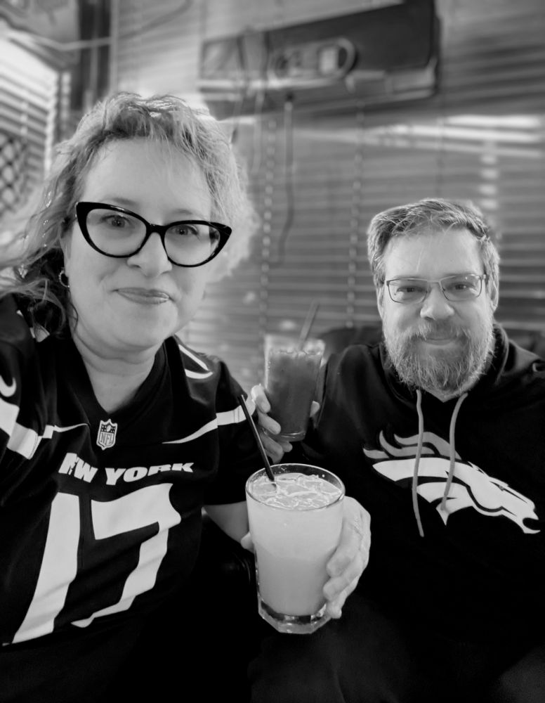 Bedhead be damned, Rickee and Jason made it for the Jets vs Broncos! The black and white photo shows a woman with cat-eye glasses in. Jets jersey holding a drink sitting next to a man with a beard holding a drink in front of horizontal blinds