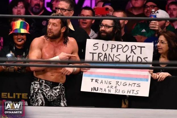 Kenny Omega, a shirtless man with curly hair, stands next to and points to a sign that says SUPPORT TRANS KIDS - TRANS RIGHTS ARE HUMAN RIGHTS