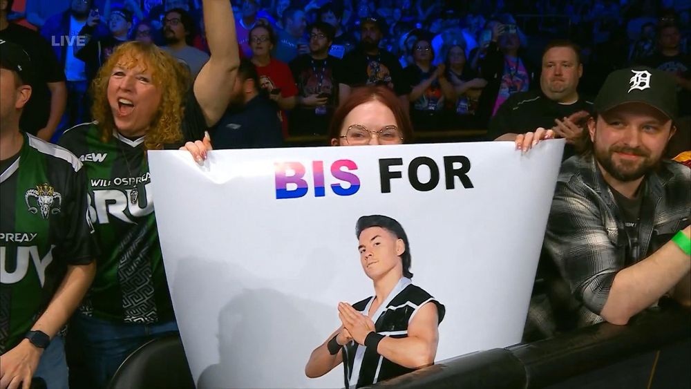 A person with dark hair and glasses peeps over a white sign that says BIS FOR [photo of Speedball Mike Bailey, a person with a dark mullet holding his hands together in a martial arts pose]