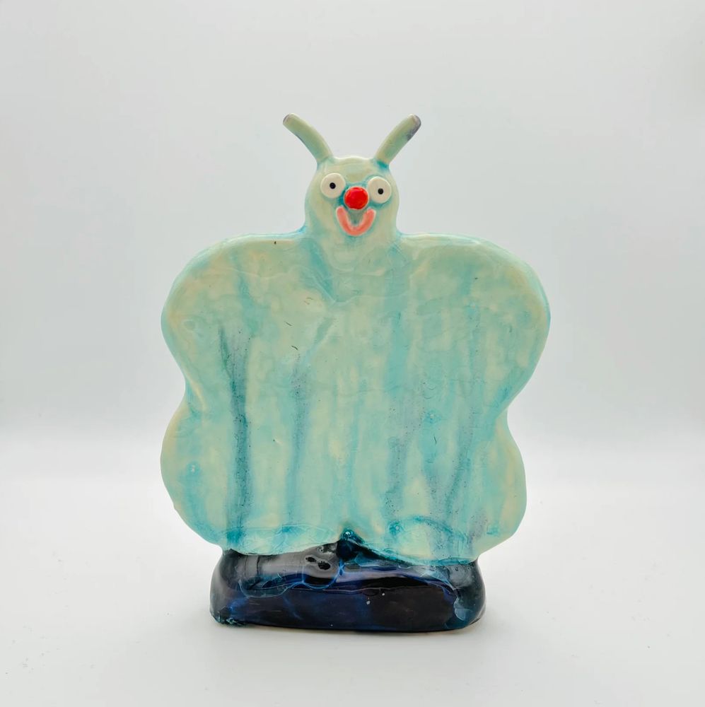 Photo of a ceramic ornament against a light grey background. The ornament is a flat cartoon butterfly. It is painted light blue and has small black and white eyes, a round red nose and smiling pink mouth. The stand is dark blue.  