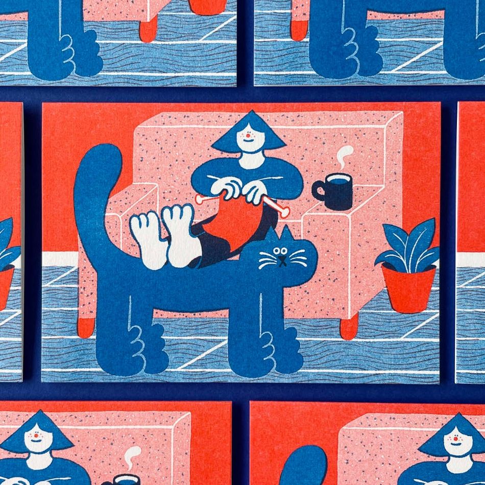 Photo of some greetings cards on a dark blue background. The cards are risograph printed in red and blue ink. They feature an illustration of a person sitting on a sofa knitting. They have a mug of hot drink and are resting their feet on the back of a large cat. 