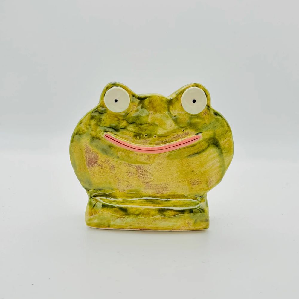 Photo of a ceramic ornament against a light grey background. The ornament is a flat cartoon frog face on a stand. It is painted in a variety of greens and has a wide pink smiling mouth, big round black and white eyes and little nostrils. 