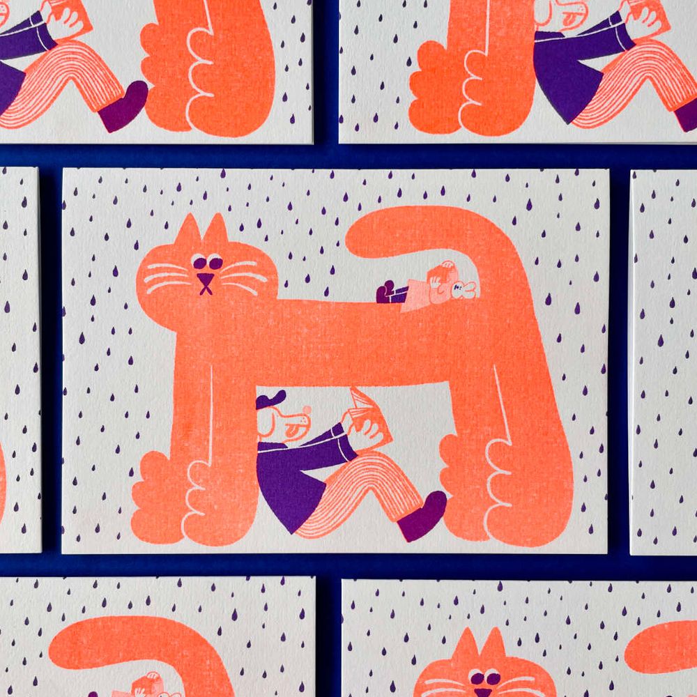 Photo of some riso-printed greetings cards laid flat on a dark blue background. The card has an illustration on the front of a large cat standing as rain falls around it. There is a dog person wearing human clothing, sitting under the cat and reading a book. Lying on top of the cat but under the tail is a mouse person in a dress reading a book. The colours are fluorescent orange and purple. 
