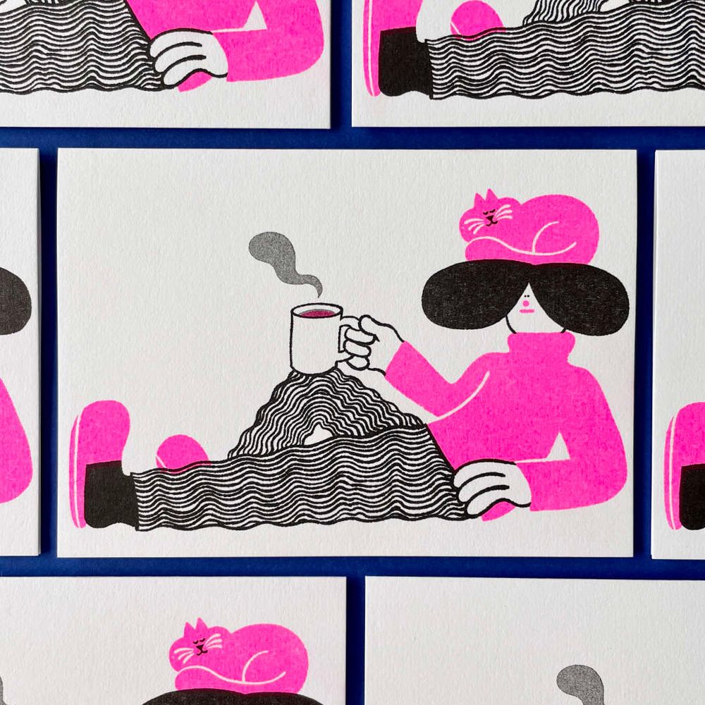 Photo of some riso-printed greetings cards laid flat on a dark blue background. The card has an illustration on the front of a person reclining with a mug of tea resting on their raised knee. There is a cat sleeping on the person's head. The colours are fluorescent pink and black on white paper. 