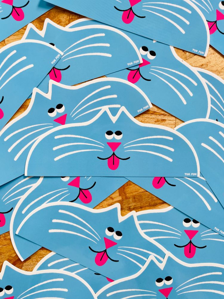 Photo of some stickers spread out on a table. The design is an illustration of a cat's face. It is looking upwards and its tongue is sticking out. The design is die-cut and is drawn in light blue, pink, black and white. 
