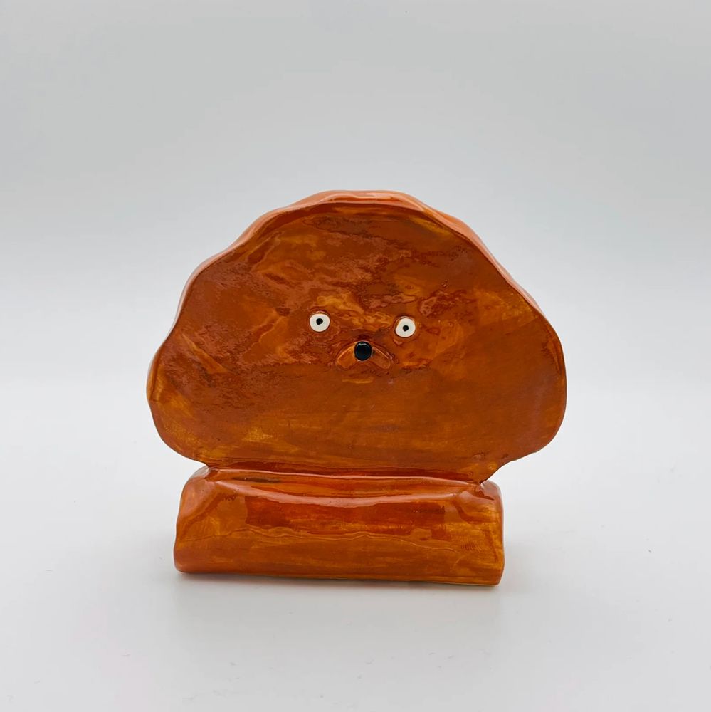 Photo of a ceramic ornament against a light grey background. The ornament is a flat cartoon dog face on a stand. It is painted brown and has small black and white eyes and a black nose. 