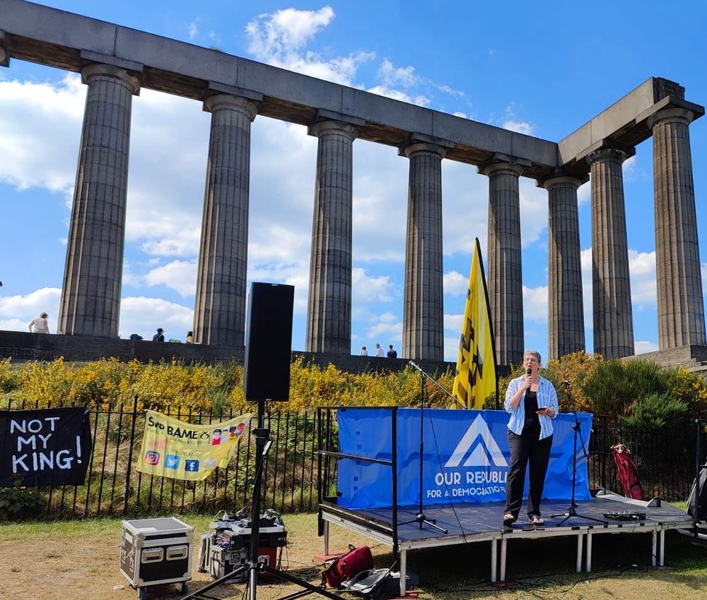 Maggie speaking at a microphone on Calton Hill. There’s blue sky and sunshine behind her, and posters saying “Not my king” next to the stage