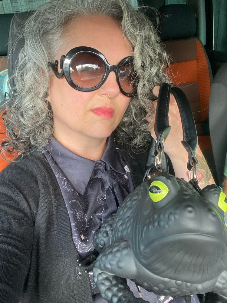 Een selfie van mij, een vrouwelijk uitziend persoon. Ik heb grijs krullend haar, een grote zwarte zonnebril, een zwart jurkje met dasje en daar een zwart vest overheen. Ik houd een tas omhoog in de vorm van een zwarte kikker 