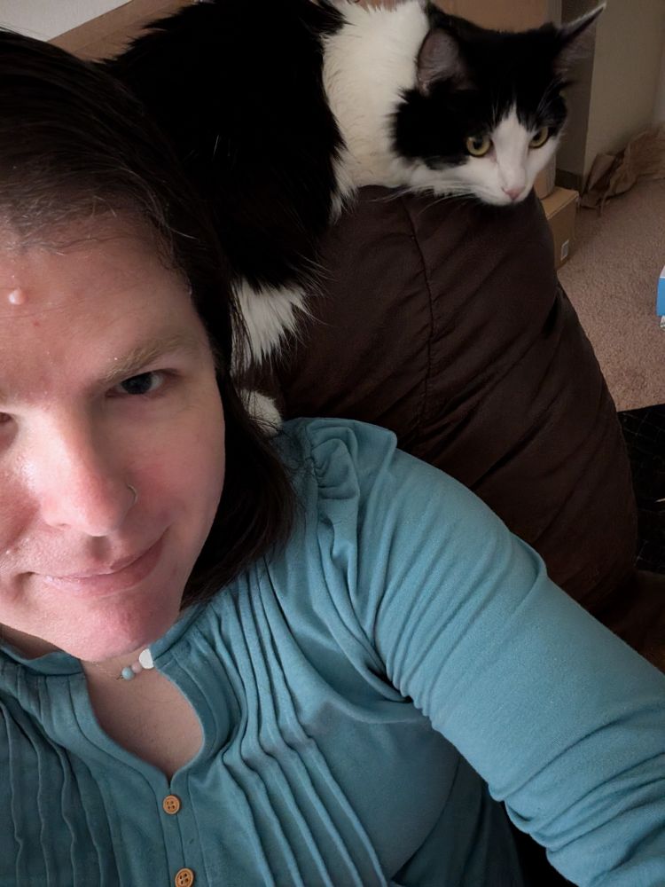 A person wearing a green blouse sitting in a brown chair with a black and white cat sitting above her