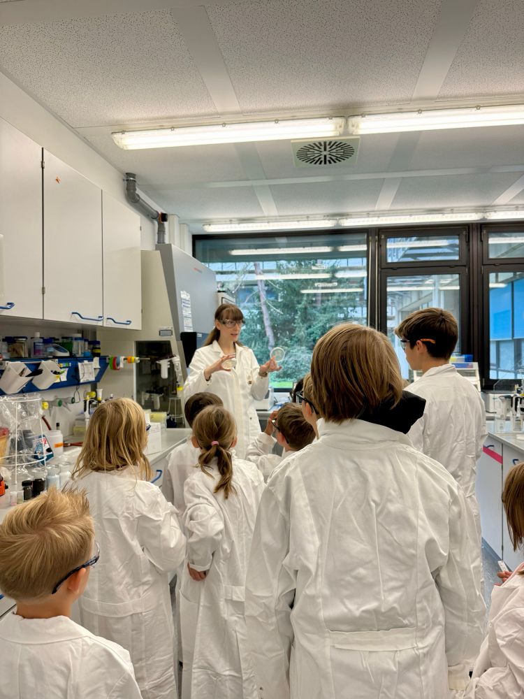 Photo of a lab tour. Kids are wearing lab coats and glasses and Sophie is showing agar plates