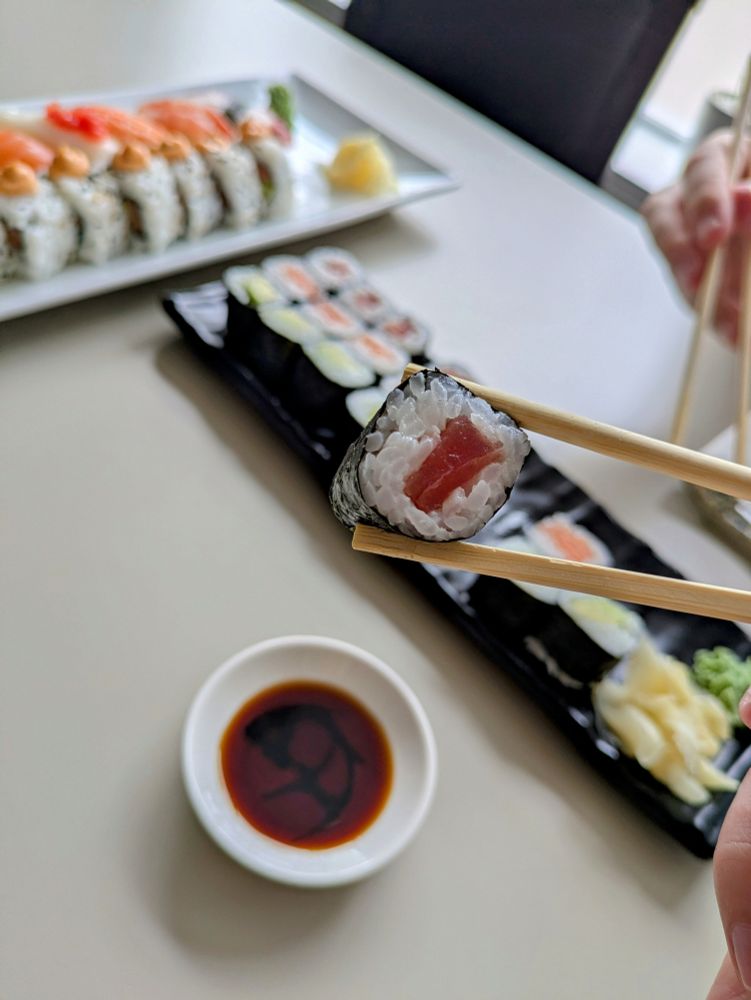 A photograph of a platter of sushi - tuna, salmon and avocado hosomaki and nigiri. One of the hosomaki rolls is being held up to the camera with a pair of chopsticks.