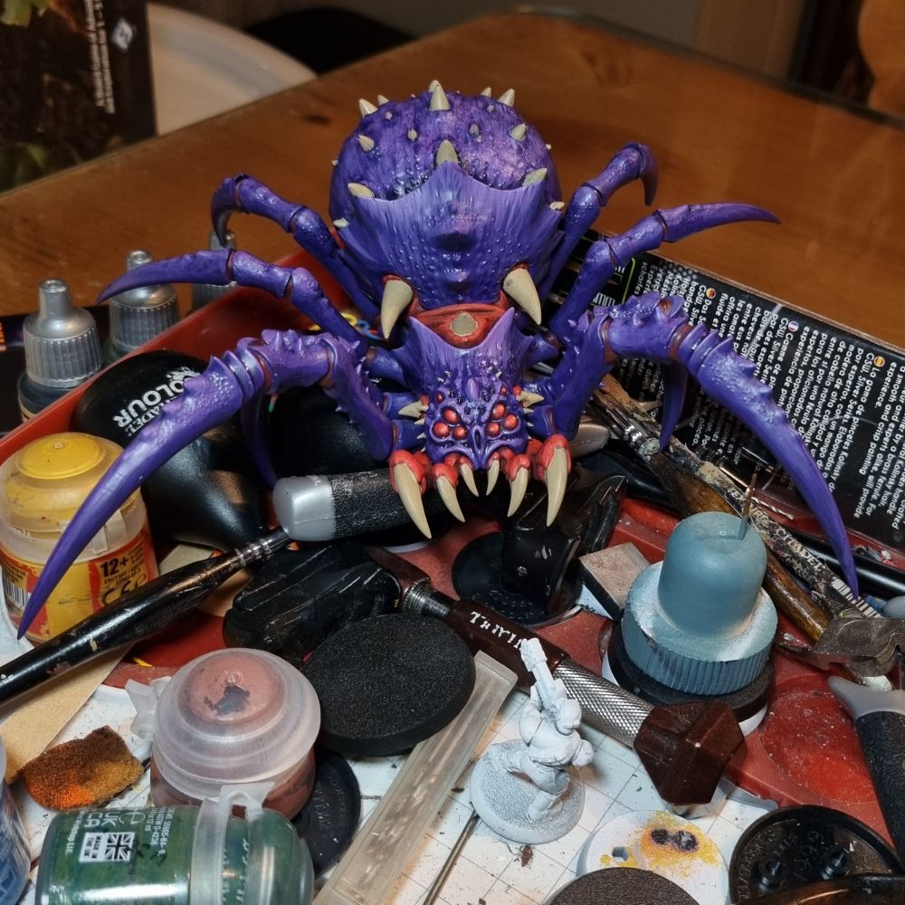 An unpainted catachan jungle fighter aiming a flamer in the mouth of a large purple spider mini.