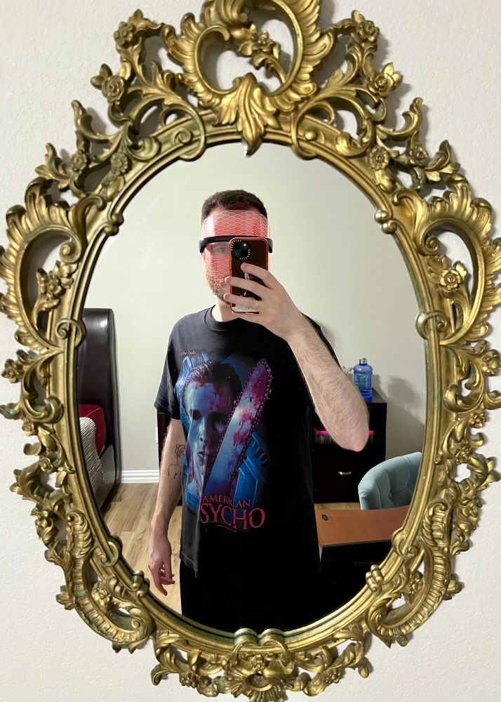 Mirror selfie of the user wearing an American Psycho t-shirt while doing their skincare routine wearing a red LED light therapy mask
