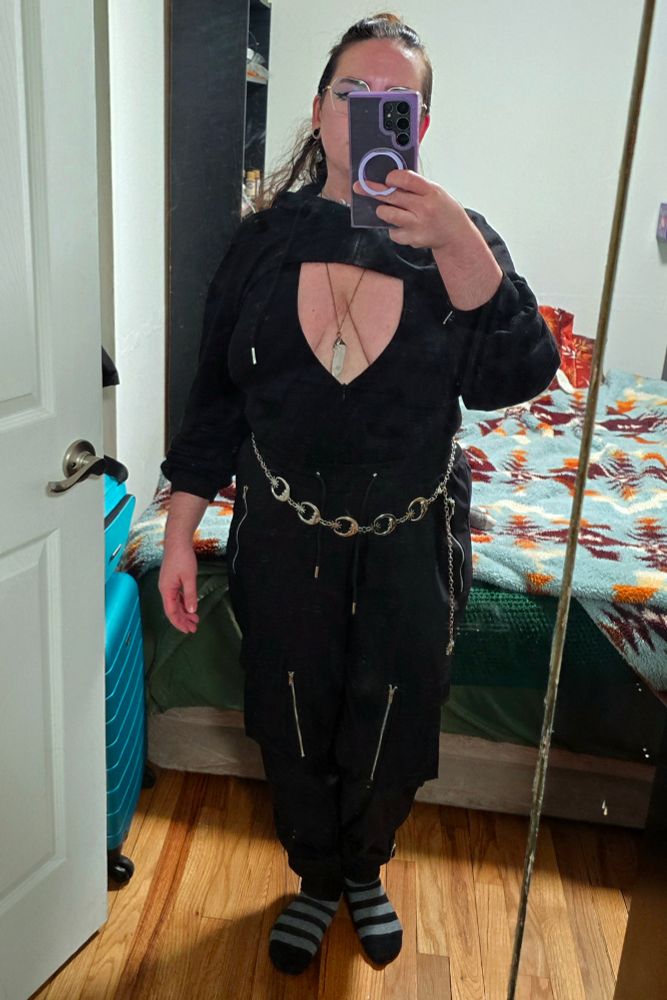 Mirror selfie of nonbinary FTM femboy Stellaris in a plunge neck tight shirt with a micro crop top hoodie, and goth cargo pants with lots of zippers. They accessorize with a silver moon and star chain belt, a long gunmetal and angel aura quartz necklace that dips between their breasts, a silver moon phase choker, and moon moth black and silver dangle tunnels. Their brunette hair is half up in a bun and theyre growing out their side shave.