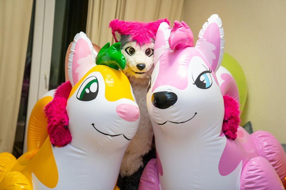 Lupe standing between two inflatable huskies - a yellow one with green eyes, green hair, pink ears and pink nose, and a pink one with light blue eyes, pink ears, dark pink hair and a black nose