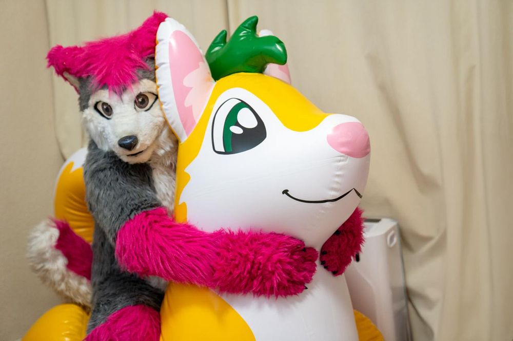 Lupe hugging a yellow inflatable husky with white front, pink nose and ears, and a green splash of hair - background is a cream curtain 