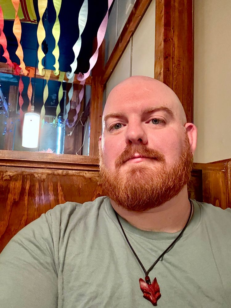 A bearded, bald man with a red fox-shaped pendant necklace poses for a selfie in a wooden booth. Rainbow streamers hang in twisted strands from the ceiling above, adding a festive and colorful touch to the background. The lighting is warm and soft.