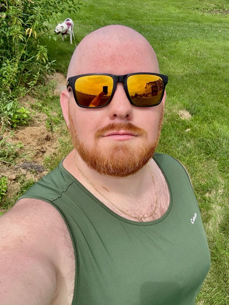 I’m outside on a grassy lawn, wearing a green tank top, gold chain, and reflective gold lense black rimed sunglasses. I’m looking at the camera with a slight smile. My phone is visible in the reflection of my sunglasses. In the background, my white dog in a pink harness is walking through the grass near some bushes.