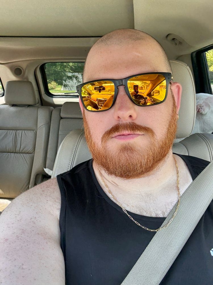 I’m sitting in the driver’s seat of my car in the drive trough. I have a shaved head and ginger beard. I am wearing a black tank top, gold chain, and reflective yellow sunglasses. The interior of the car is light-colored, and part of my dog’s head is visible behind me on the right.