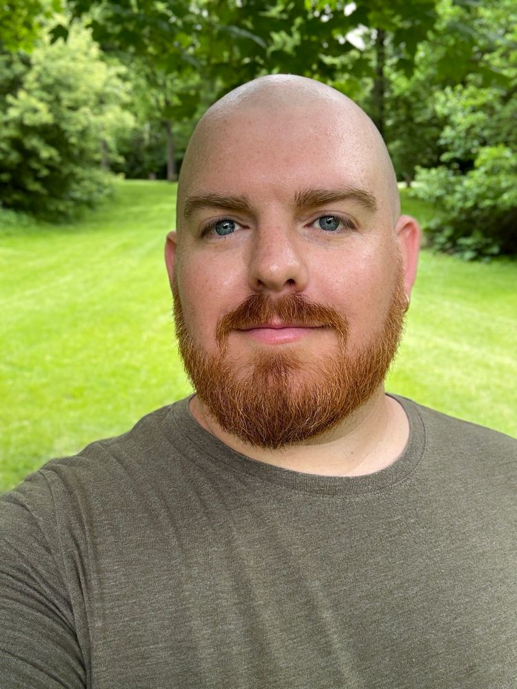 Alt text: A bearded man with a shaved head and red hair is taking a selfie outdoors. He is wearing an olive green t-shirt and standing on a lush green lawn surrounded by leafy trees. The background features a pathway of mowed grass leading into a wooded area, creating a serene, natural setting.