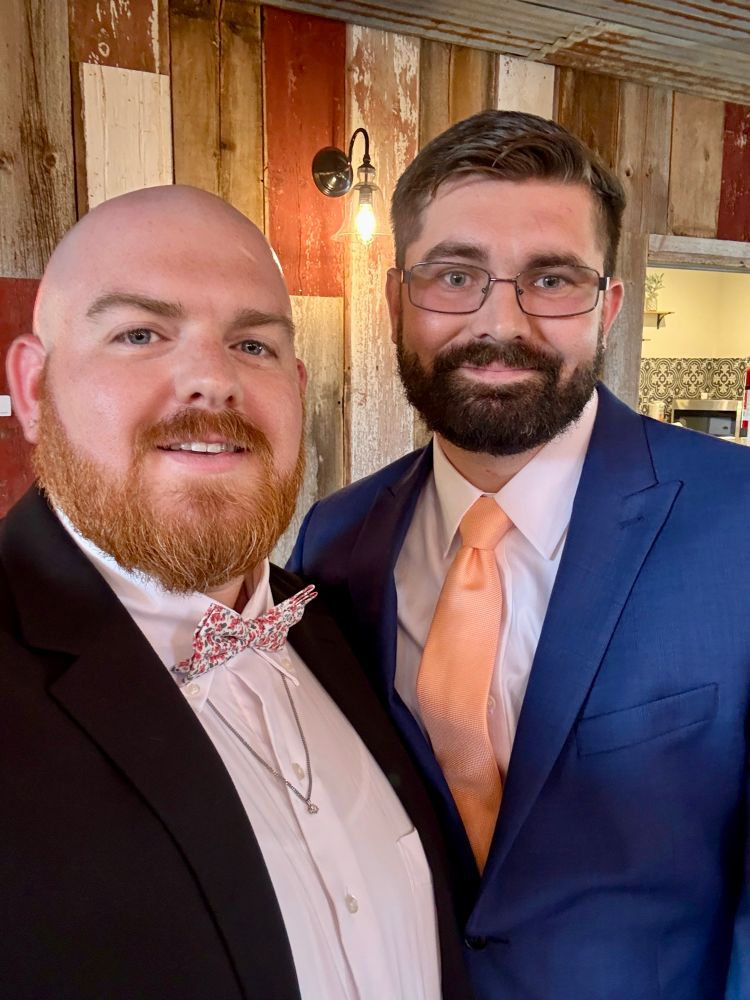 Two men dressed in formal attire pose for a selfie in front of a rustic wood-paneled wall with a vintage-style light fixture. The man on the left has a bald head, red beard, floral bow tie, and a black suit jacket over a white shirt with a necklace. The man on the right has short dark hair, glasses, a neatly trimmed beard, and is wearing a blue suit with a peach-colored tie and white shirt. Both are smiling warmly.