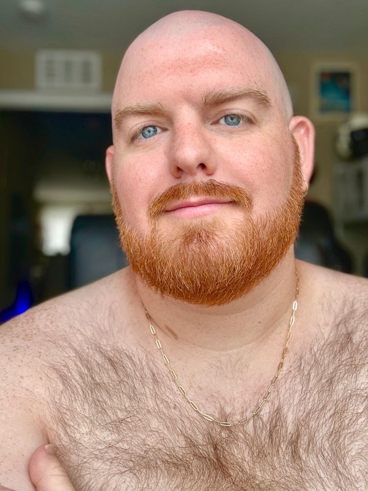I’m indoors, looking at the camera with a relaxed expression. I’m shirtless, showing my chest hair and wearing a gold chain. The lighting is soft, and the background includes a doorway and some blurred room details.