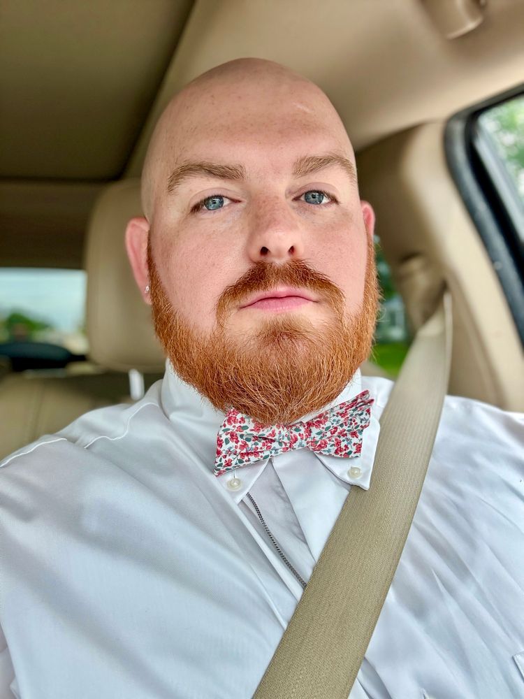 A bald man with a red beard sits in the driver’s seat of a car, wearing a white dress shirt, a red and white floral bow tie, and a seatbelt across his chest. His expression is smirked, and natural light brightens his face