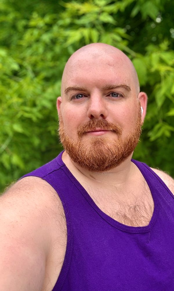 A bearded bald man wearing a purple tank top is taking a selfie outdoors. He has light skin, reddish facial hair, and is wearing a white earbud. Behind him is a lush background of green leafy trees, indicating a warm, natural setting.