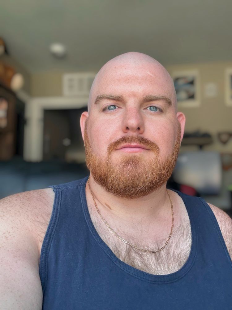 Indoors, shaved head bald ginger bearded, wearing a navy blue tank top and a gold chain while showing chest hair. I’m looking directly at the camera with a neutral expression. The background is softly blurred, showing a doorway, shelves, and framed pictures on the wall.