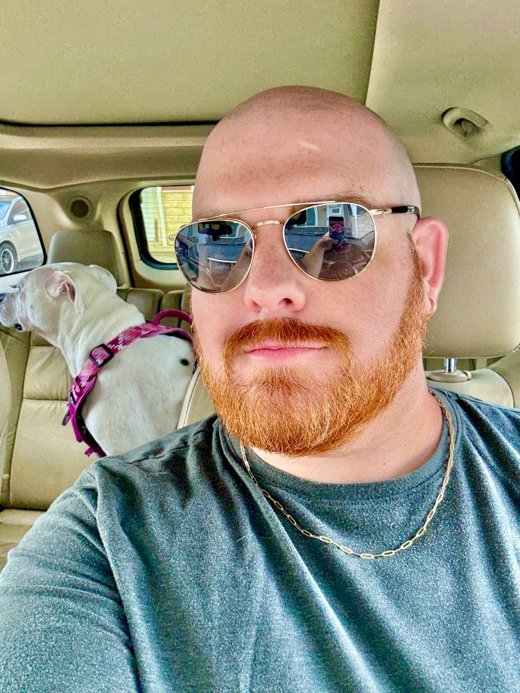 A selfie of myself a bald man with a red beard and sunglasses, sitting in the front seat of a car wearing a dark grey shirt and a gold chain necklace. In the back seat, a white dog wearing a pink harness looks out the window on the opposite side of the car. The interior of the vehicle is beige, and sunlight streams in through the windows.