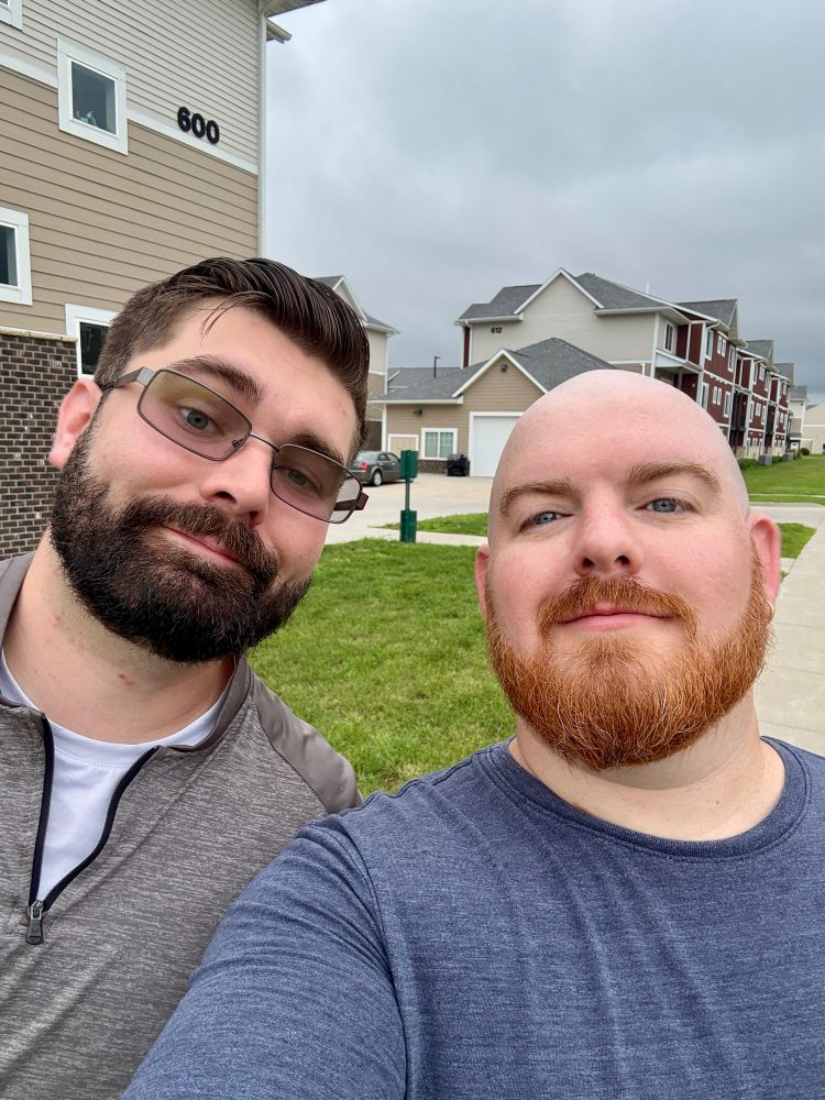 Two men take a selfie outside on a cloudy day in a suburban neighborhood. The man on the left has dark hair, glasses, a full beard, and is wearing a gray quarter-zip pullover. The man on the right is bald with a red beard and is wearing a blue crewneck shirt. Behind them are multi-story residential buildings with a neatly maintained lawn.