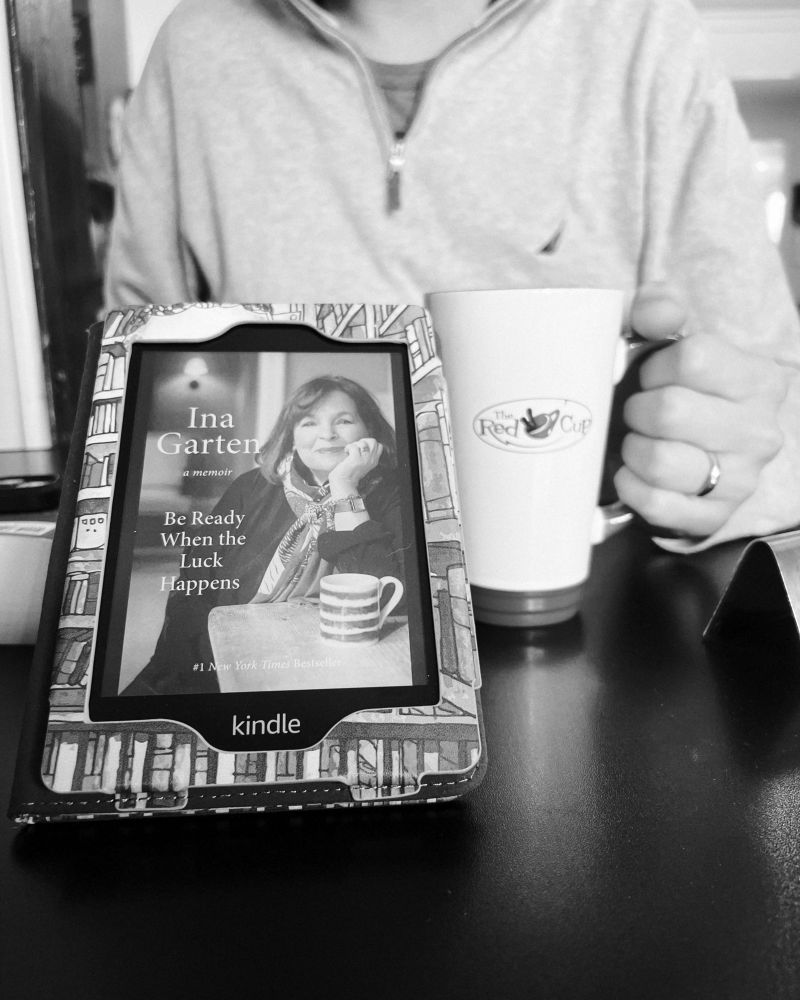 A black-and-white image of a kindle displaying the cover of Be Ready When the Luck Happens by Ina Garten. In the background, a person holds a coffee cup