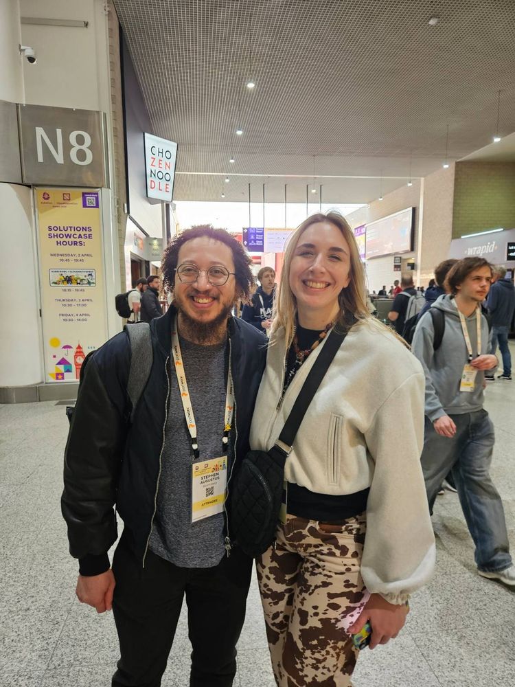 Megan and I, doing the hallway track at KubeCon London. She's wearing cool pants with a cow print on them.