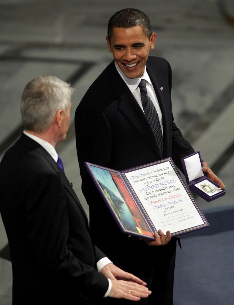Barack Obama standing with a white haired gentleman, posing with a certificate and medal for the 2009 Nobel Peace Prize