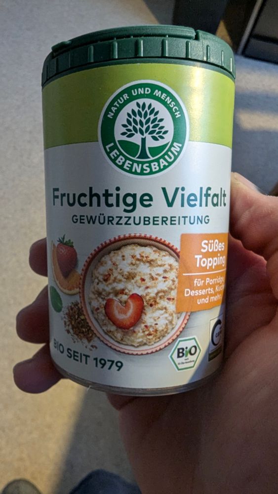 Das Bild zeigt eine Hand, die ein zylindrisches, grünes und beigefarbenes Produkt in einer Kunststoffverpackung hält. Es handelt sich um ein Lebensmittelprodukt der Marke „Lebensbaum“.
Auf der Verpackung sind folgende Texte und Bilder zu sehen:
 * Ein runder, grüner Aufkleber mit einem Baum in der Mitte und den Worten „Natur und Mensch“ und „Lebensbaum“.
 * In großen Buchstaben steht „Fruchtige Vielfalt“ gefolgt von „Gewürzzubereitung“ in kleinerer Schrift.
 * Daneben ist eine Schale mit Porridge abgebildet, der mit Erdbeeren garniert ist. Eine der Erdbeerscheiben ist in Form eines Herzens geschnitten.
 * Ein weiteres orangefarbenes Feld weist auf ein „Süßes Topping“ hin, das für „Porridge, Desserts, Kuchen und mehr“ geeignet ist.
 * Links von der Schale ist eine halbe Erdbeere und eine Orangenscheibe zu sehen.
 * Unten links steht „BIO SEIT 1979“.
 * Rechts unten ist ein Bio-Siegel mit dem Text „BIO“ und das „C“ in einem Kreis, was auf eine Zertifizierung hinweist.
Der Deckel der Verpackung ist dunkelgrün. 
