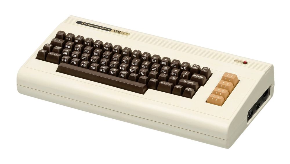 A Commodore VIC-20 home computer from the early 1980s. The machine has a cream-colored plastic casing with a built-in keyboard featuring brown keys with white lettering. The function keys on the right side are beige. The “Commodore VIC-20” logo is printed in black and gold on the top left. The keyboard includes PETSCII graphic symbols, color control keys, and a layout similar to later Commodore models. A red LED power indicator is positioned in the top right corner. On the right side of the machine, there are various input/output ports for peripherals. The VIC-20 was known as one of the first affordable color home computers, popular for gaming and programming.
