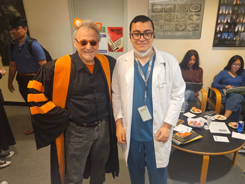 A professor wearing black academic regalia with orange stripes, tiger ears, and orange sunglasses, poses with a student in a white lab coat and blue scrubs at a Halloween gathering in the history department.