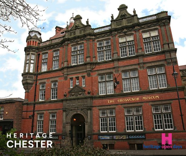 A photograph of the Grosvenor Museum, a Victorian red brick building with ornate architectural details. The three-story structure features decorative stonework, multiple rows of tall sash windows with white frames, and elaborate carved stone elements including balustrades and ornamental features on the roofline. The building has "THE GROSVENOR MUSEUM" inscribed in gold lettering on its facade. The entrance is through an arched doorway with classical stonework. Bare tree branches are visible in the foreground, and the sky shows blue with white clouds. In the bottom left corner is white text reading "HERITAGE CHESTER" and in the bottom right is the Heritage Open Days logo featuring a pink castle icon with blue and pink text.