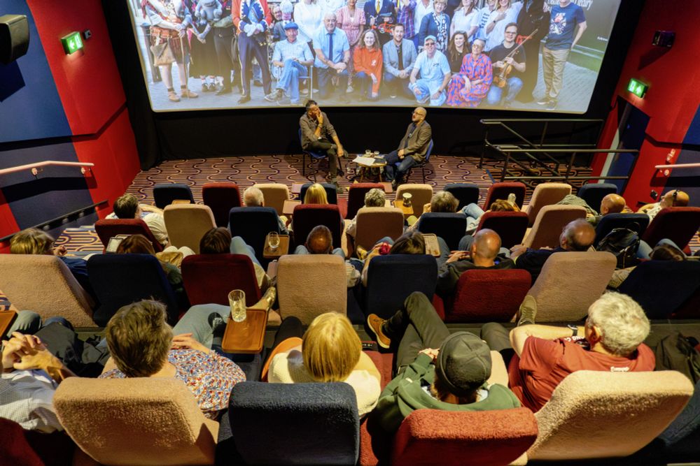Two men seated in front of a large screen, facing a large audience who are listening to them discuss events from the last twelve years. The walls are red and blue, and the seats muted pastel colours.
