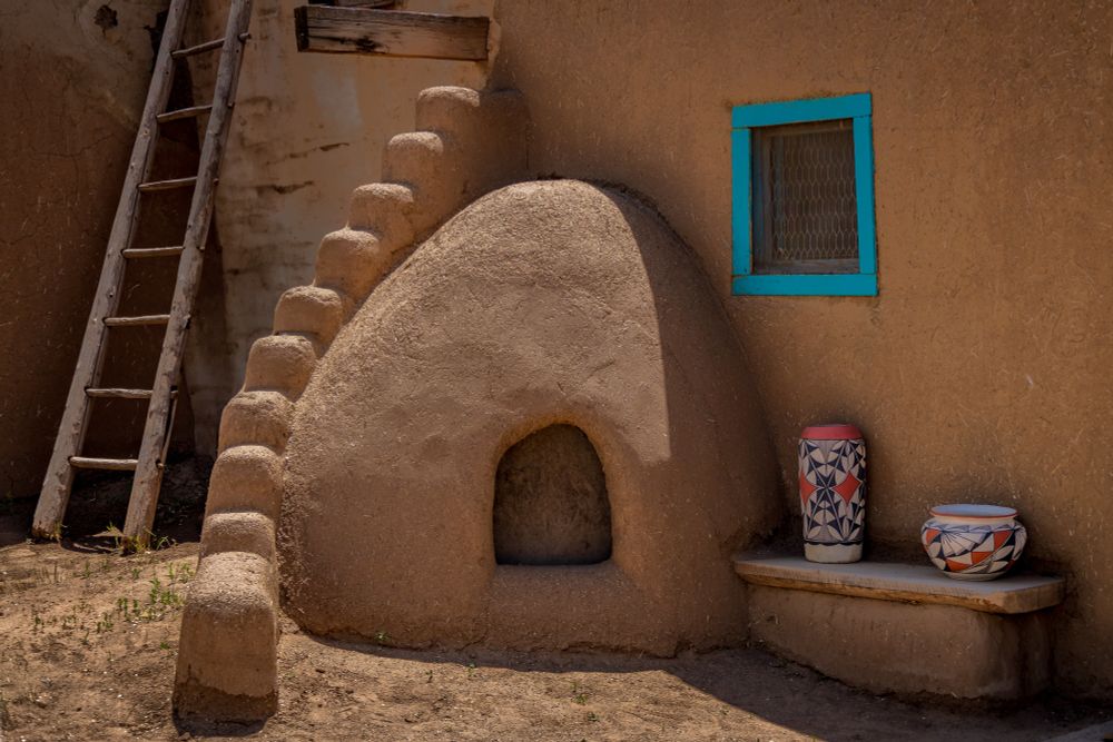 Photo of the outside of a home in Taos Pueblo, the adobe structure, featuring an adobe oven, some pots on display and a rustic ladder to the next floor