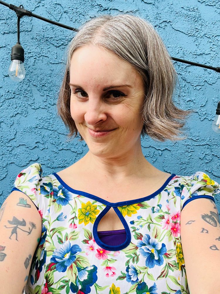 Selfie of Jen standing in front of a blue wall wearing a white dress with colourful flower pattern. 