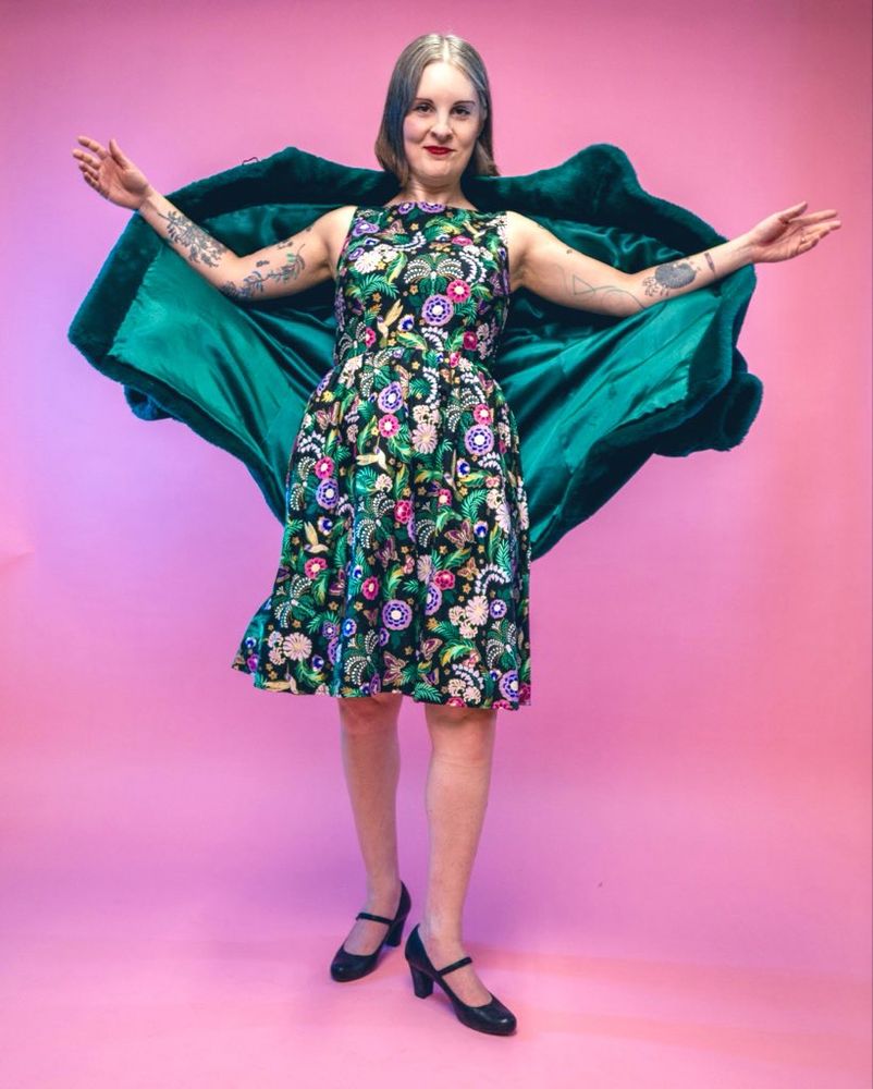 Photo of Jen from November 2024 standing in front of a pink backdrop wearing a black sleeveless dress with colourful flower pattern and flinging off a dark green faux fur coat with satin lining.