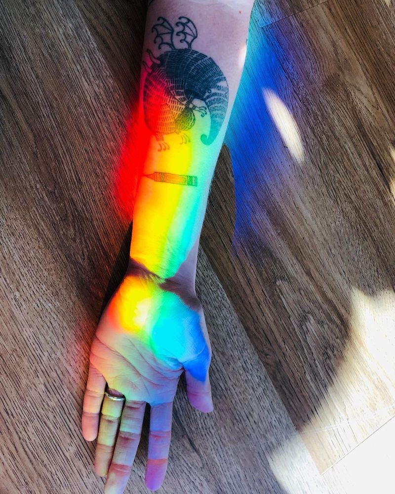 Photo of Jen’s arm on a laminate floor with tattoo of a girl with a fantastical creature named Trevor with a crayon and a rainbow (reflecting off a crystal in the window).