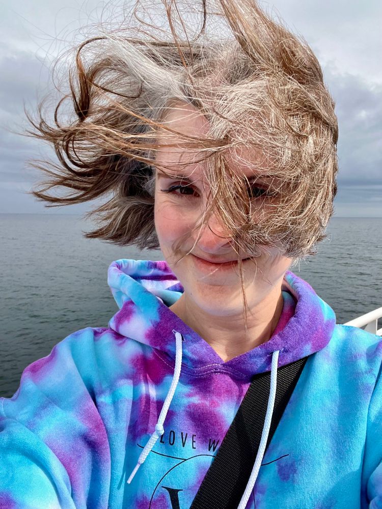 Selfie of Jen on the ferry wearing a blue and purple tie dye hoodie with windswept hair. 