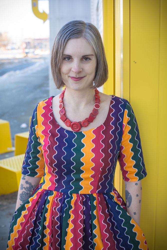 Photo of Jen from January 2022 wearing a rainbow zig zag pattern dress and red button necklace, standing next to a yellow and white building. 