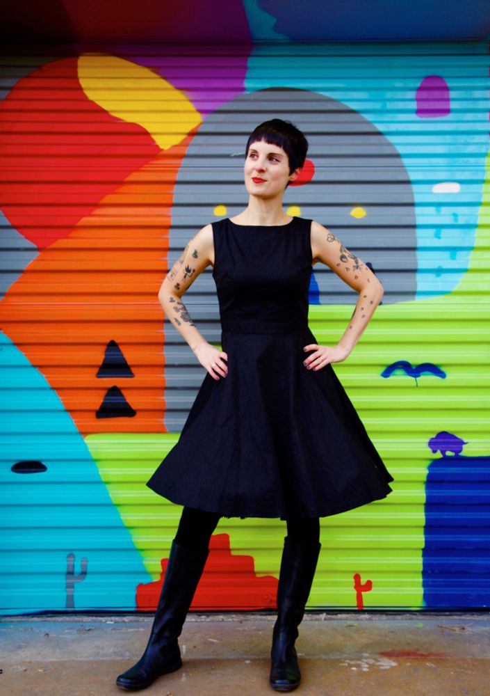 Photo of Jen from December 2017, standing against a wall painted with a multicolored mural and wearing a black dress, tights, boots and hands on hips.